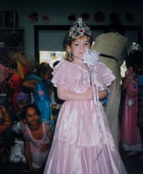 Ruth as a little girl. She's White and dressed in a pink princess dress with a crown and a wand.